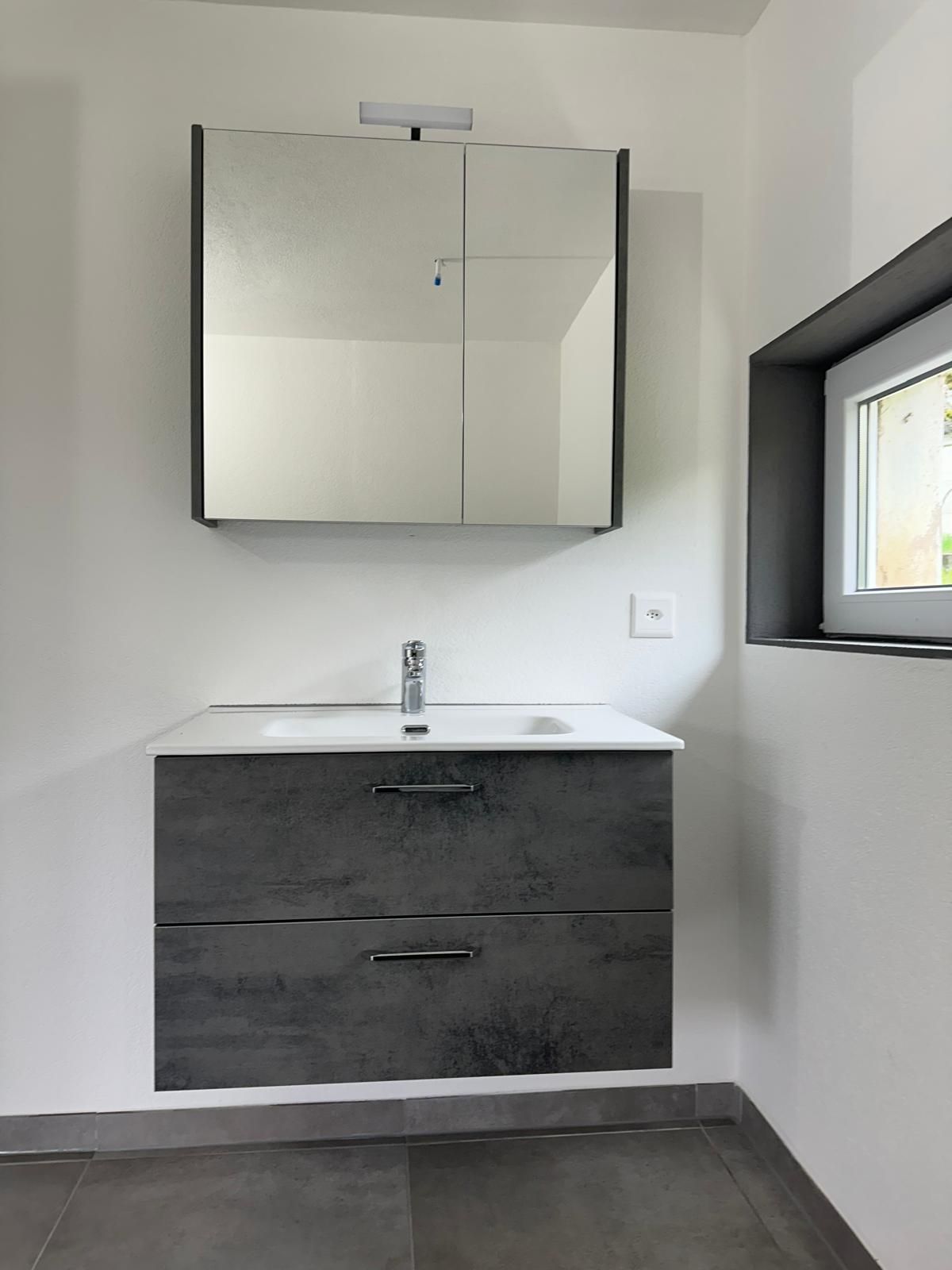 Salle de bains moderne avec murs carrelés gris, baignoire, lavabo et toilettes blancs, et miroir rétroéclairé.