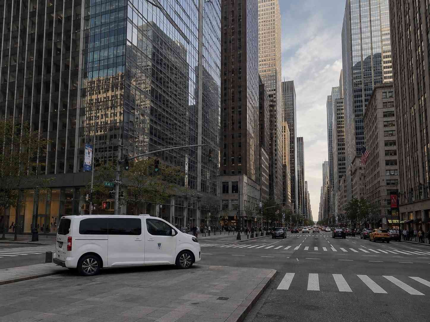 Una amplia avenida de la ciudad bordeada de altos rascacielos, con una furgoneta blanca cruzando una intersección en un día nublado.