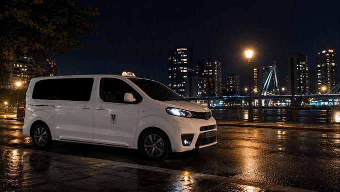 Una furgoneta blanca aparcada en una calle mojada por la noche, con las luces del horizonte de la ciudad al fondo.