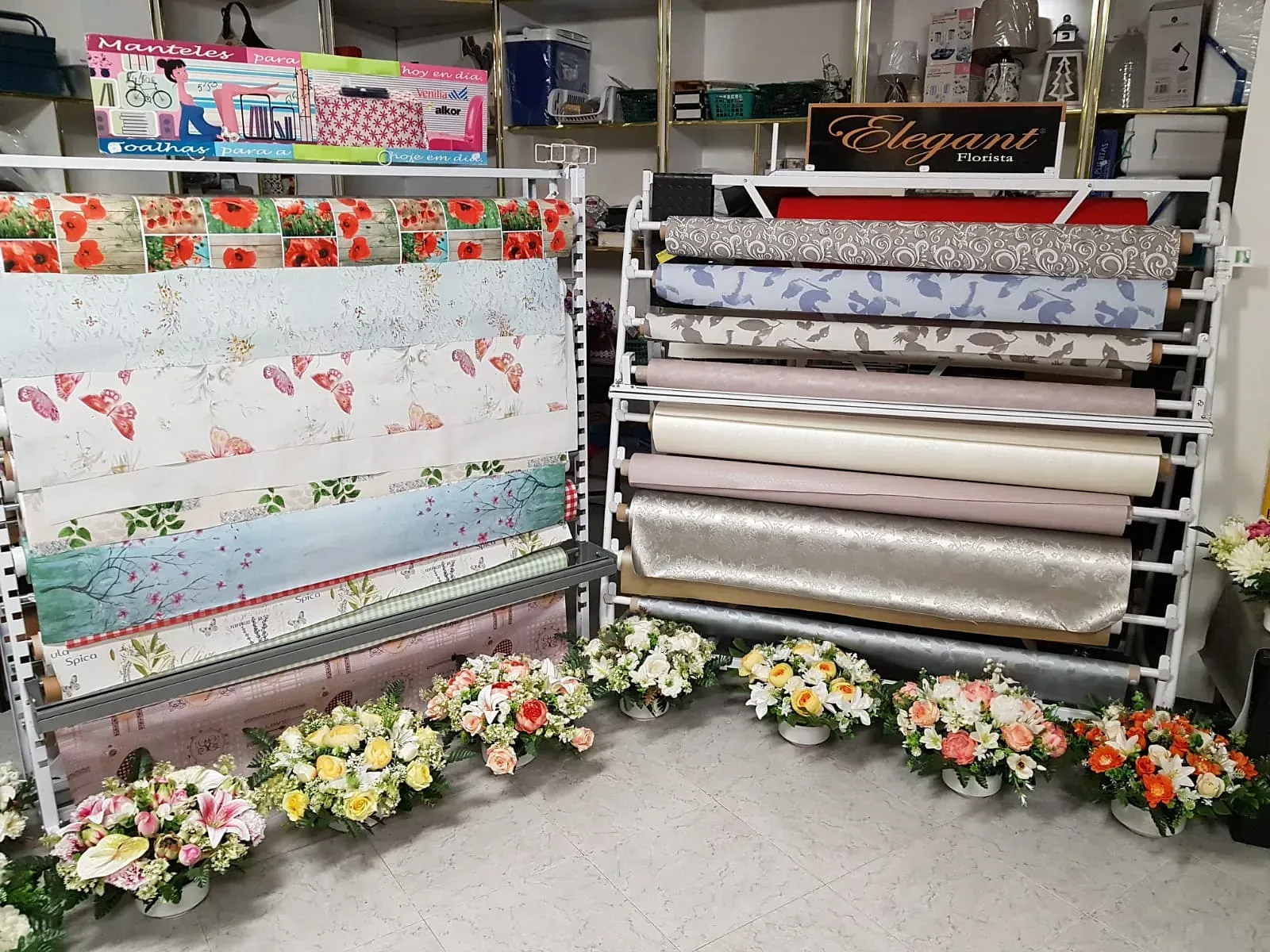 Exhibición de rollos de papel decorativo en una tienda, con estampados florales y texturas variadas. Varios ramos de flores se disponen debajo de los expositores.