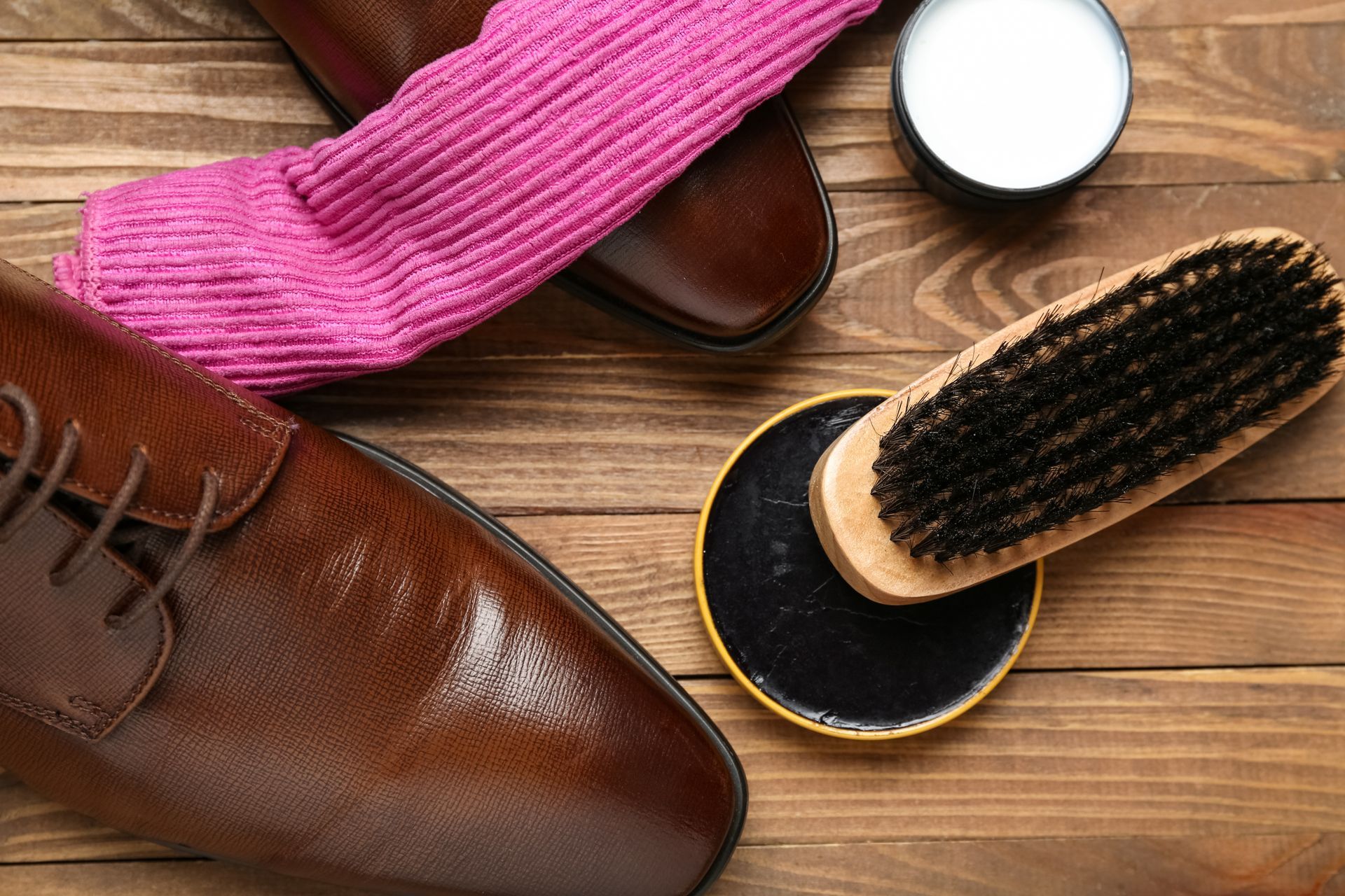 Des chaussures en cuir marron posées sur une surface en bois, avec une chaussette rose, une brosse à chaussures et des boîtes de cirage.