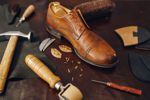 Une chaussure en cuir beige repose sur une surface sombre, entourée d'outils de cordonnerie, dont un marteau, un rouleau et de petits clous.