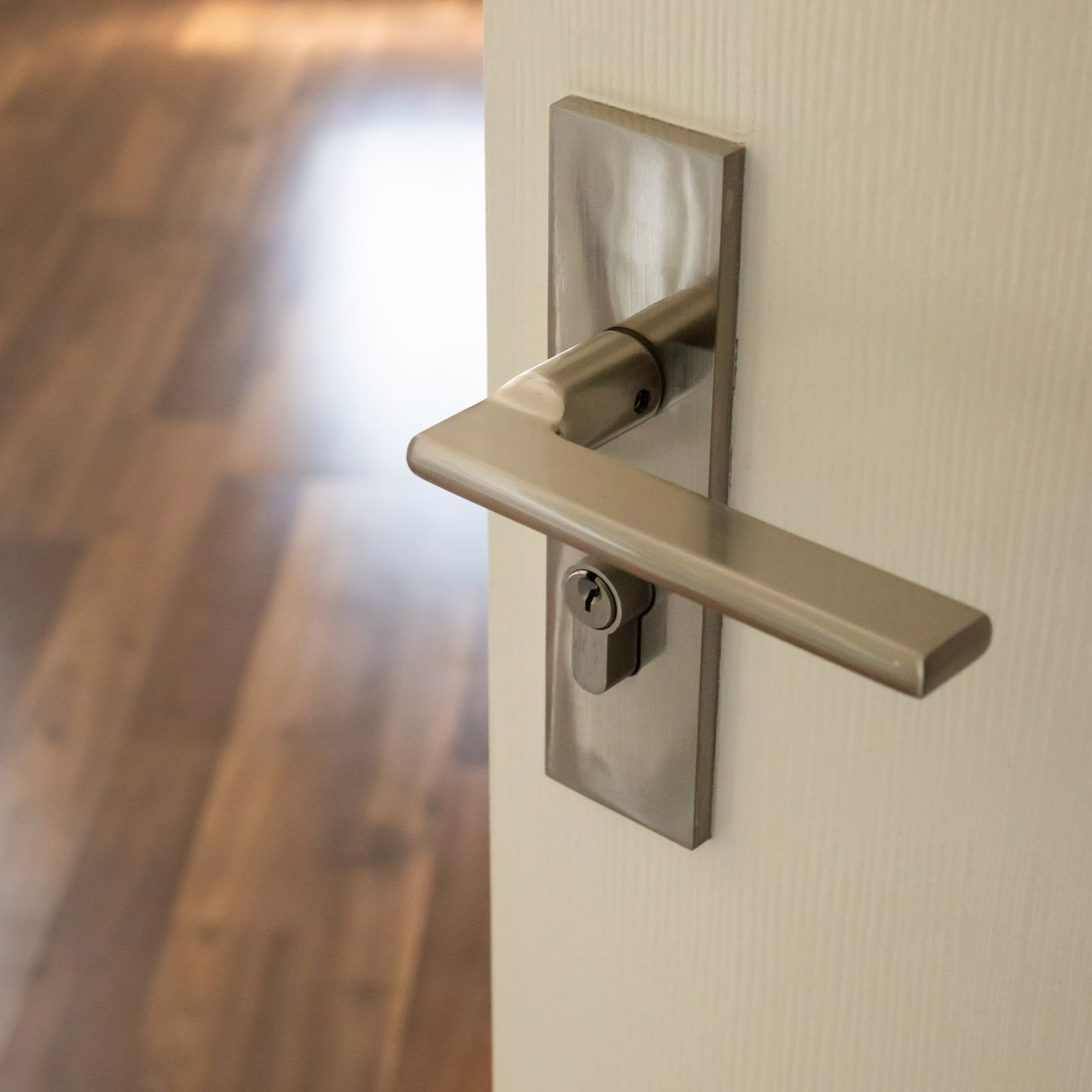 Une poignée de porte et une plaque de serrure modernes en métal brossé, montées sur une porte blanche unie, avec une pièce intérieure floue en arrière-plan.