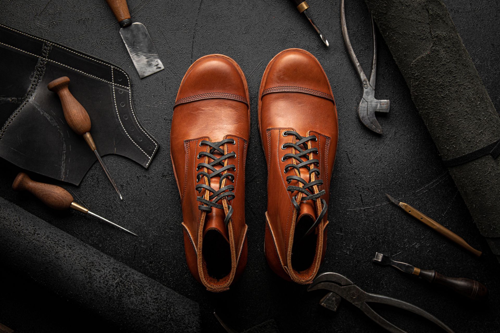 Une paire de bottes en cuir beige entourées d'outils de cordonnerie et de chutes de cuir sur une surface sombre et texturée.