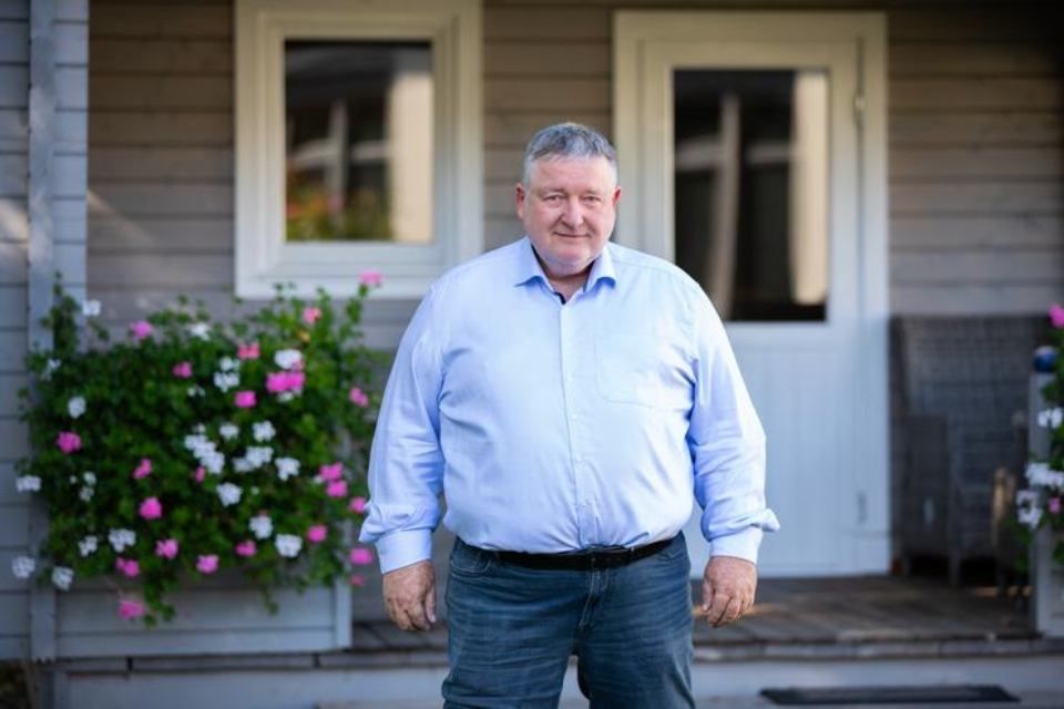 Ein Mann in einem blauen Hemd und Jeans steht vor einem Haus.