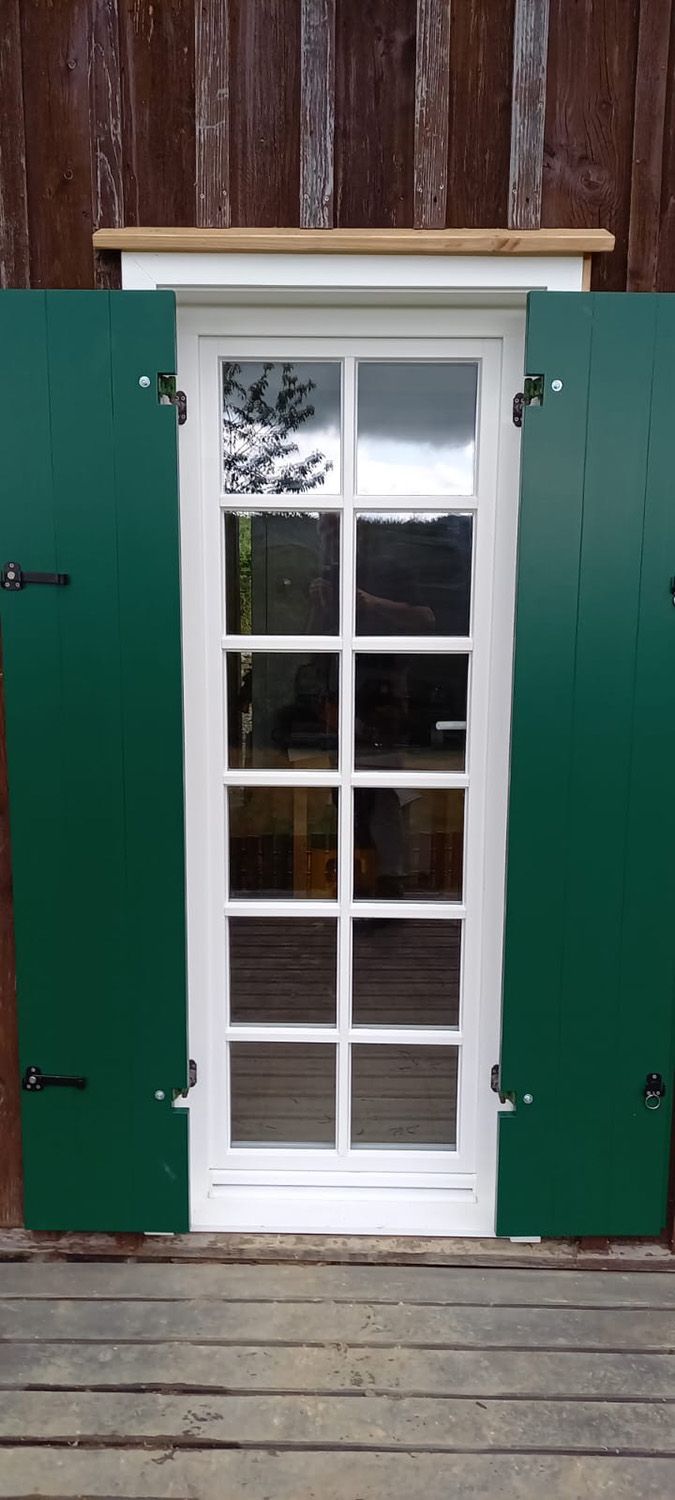 Eine weiße Tür mit grünen Fensterläden steht offen und führt auf ein Holzdeck.