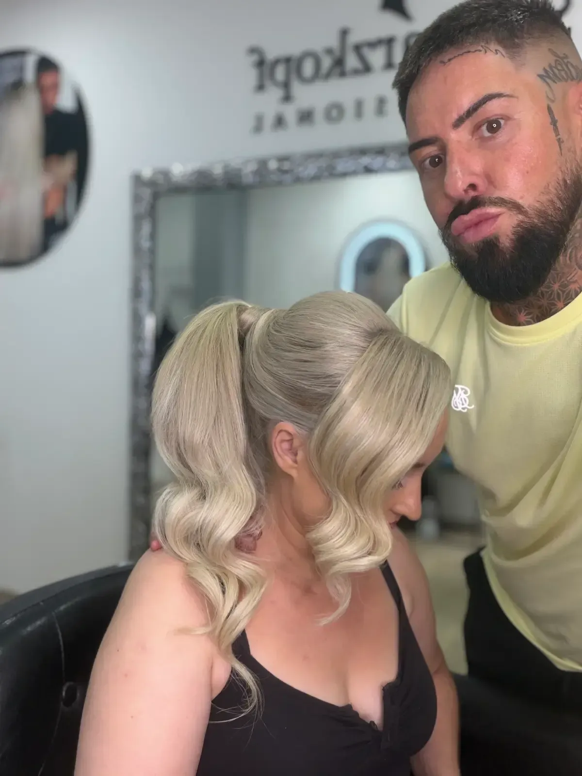 Estilista de pie junto a una clienta sentada con cabello rubio rizado en un salón de belleza.