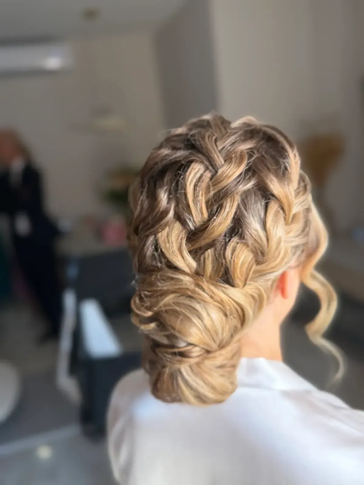 Peinado recogido con trenzas y rizos sueltos, visto desde atrás en un ambiente interior con iluminación tenue.
