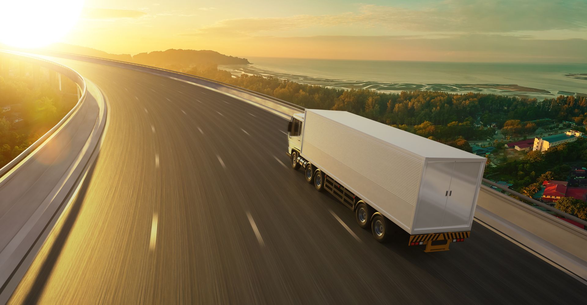 Un camion blanc roule sur une autoroute sinueuse au coucher du soleil, avec une lumière dorée et un paysage urbain au loin.