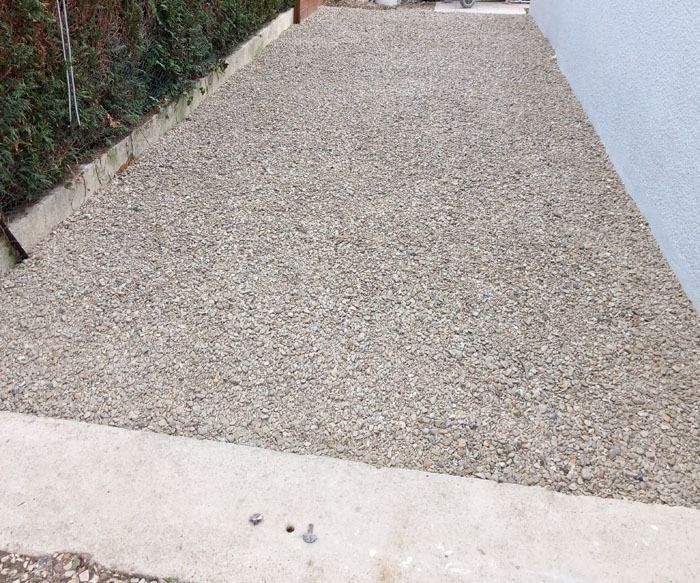 Allée en gravier entre une haie et un mur blanc, bordée de bordures en béton