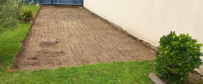 Cour en terre nue à côté d’une maison beige, bordée d’herbe et fermée au fond par un portail bleu.