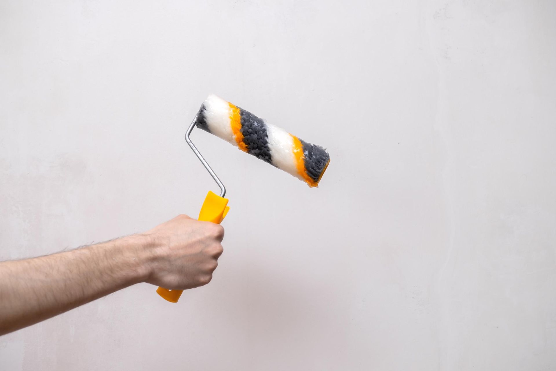 Una mano sostiene un rodillo de pintura con cerdas rayadas de color blanco, amarillo y negro contra una pared blanca lisa.