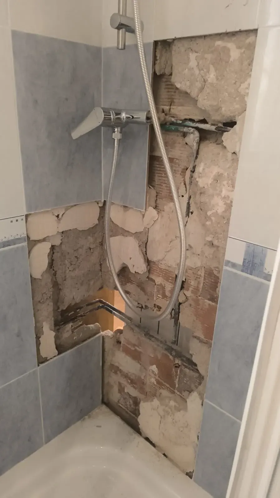 Pared de ducha dañada con yeso expuesto y azulejos rotos alrededor del cabezal de la ducha y la barra de apoyo.
