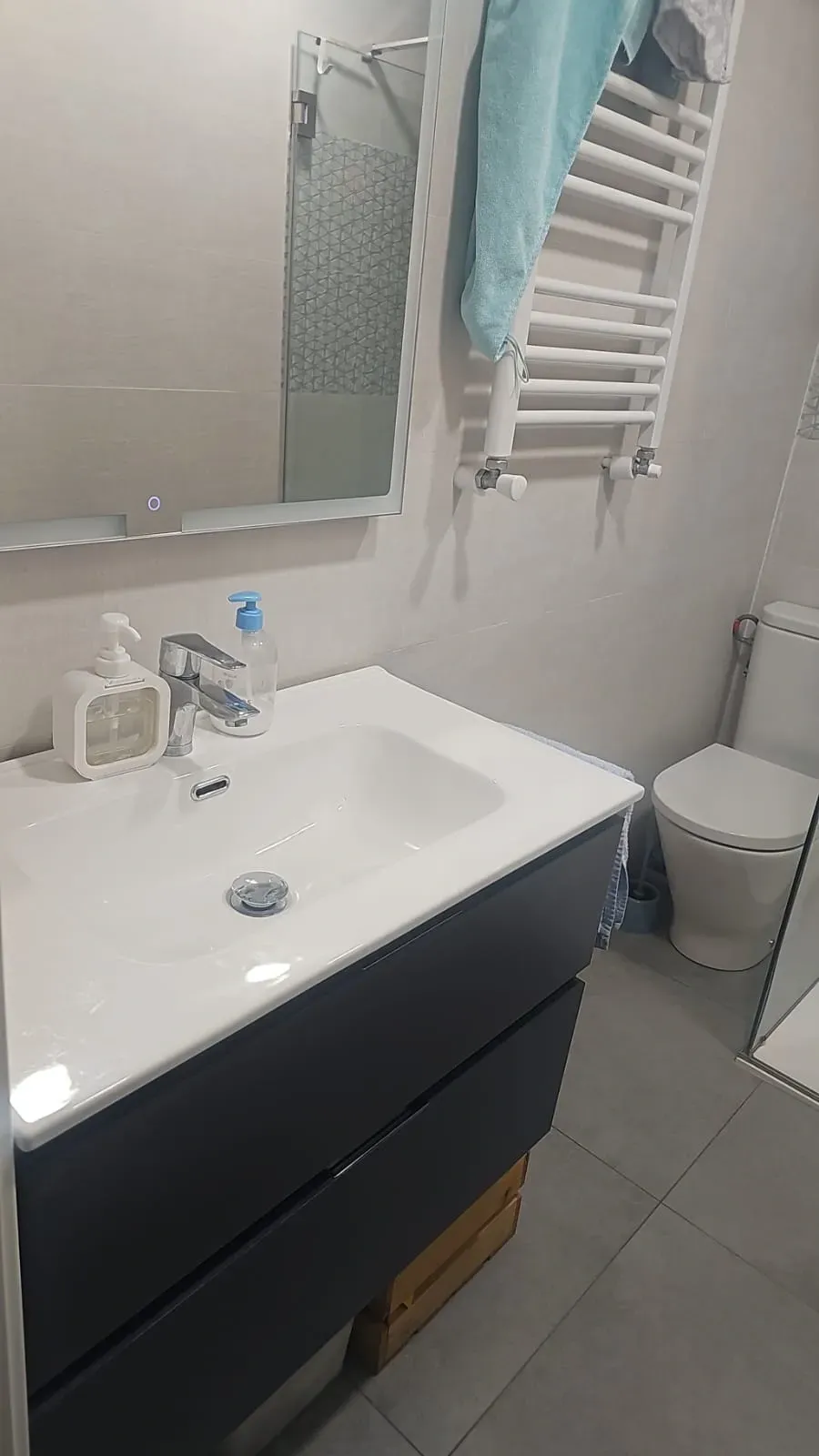 Baño pequeño con lavabo, espejo, toallero, inodoro y suelo de baldosas; una toalla cuelga junto al espejo.