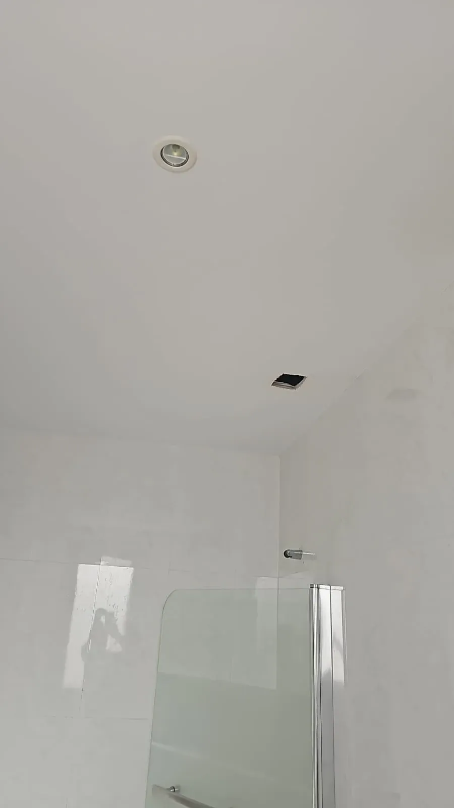 Techo blanco del baño con luz empotrada, una abertura y parte de una mampara de ducha de cristal.