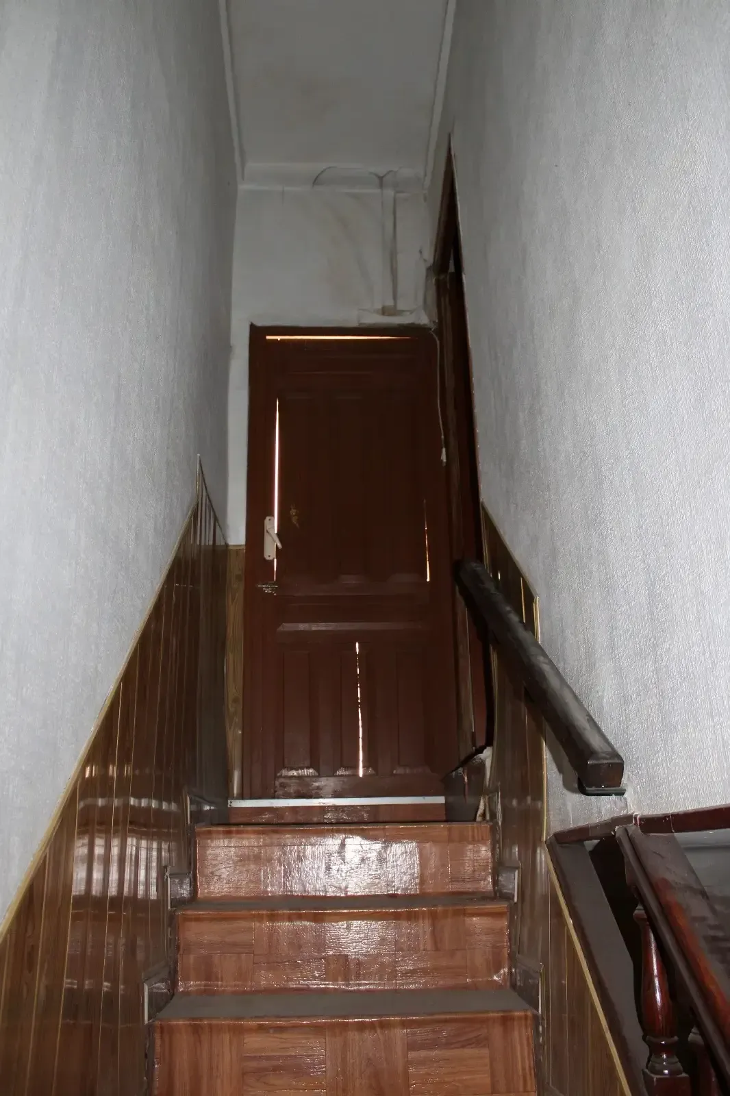 Escalera interior estrecha con escalones de madera, paredes marrones y una puerta cerrada en la parte superior.