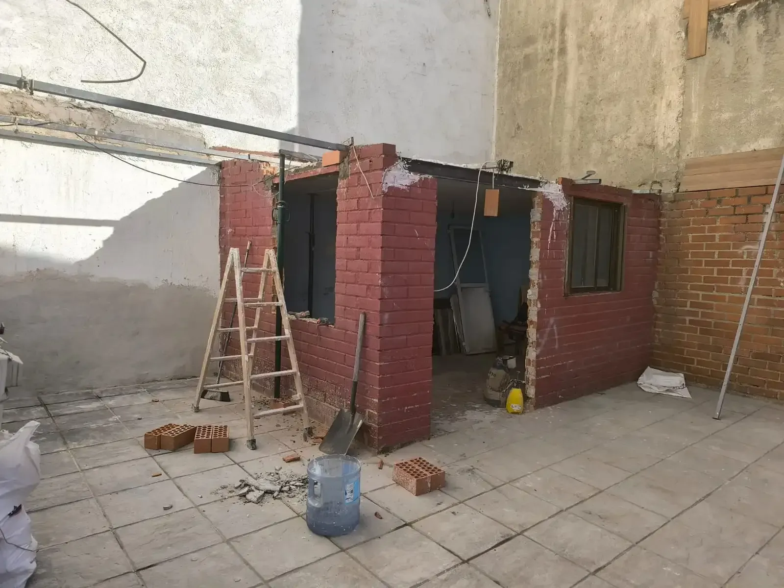 Cobertizo de ladrillo parcialmente construido en un patio, con escalera, ladrillos esparcidos y materiales de construcción.