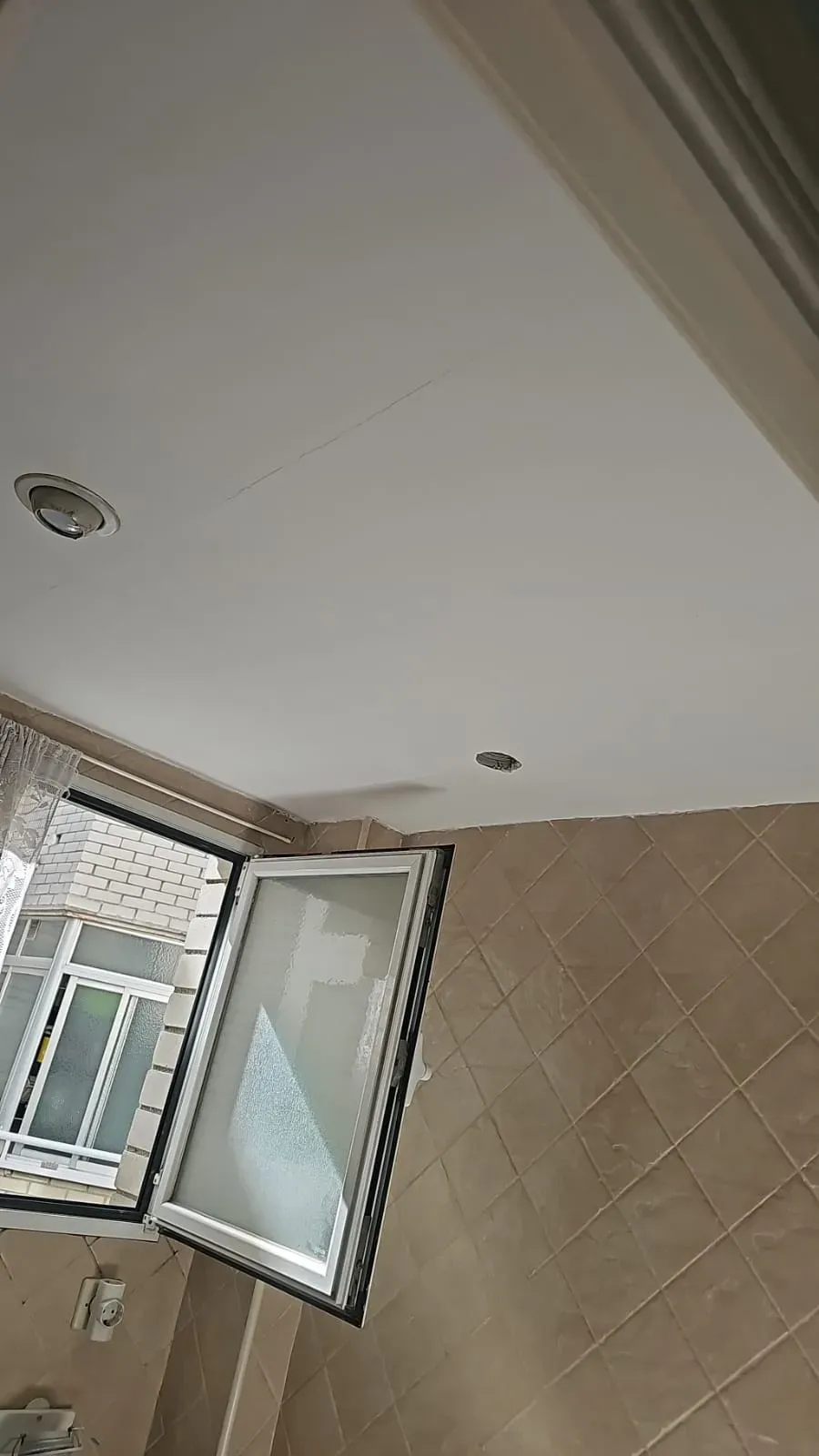 Baño con techo blanco, pared alicatada en color beige y una ventana abierta que deja entrar la luz natural.