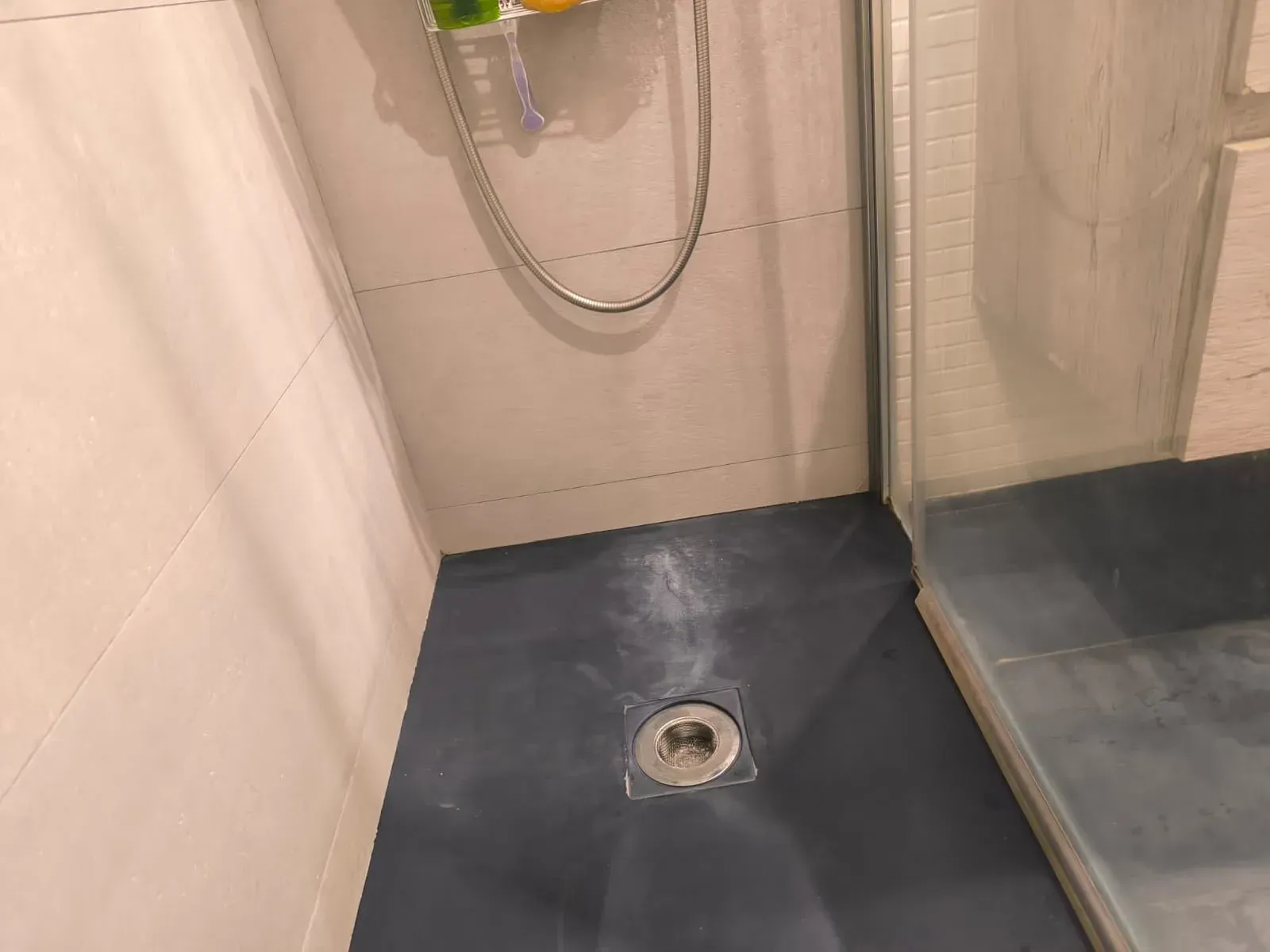 Cabina de ducha vacía con suelo oscuro, desagüe y manguera de ducha montada en la pared.