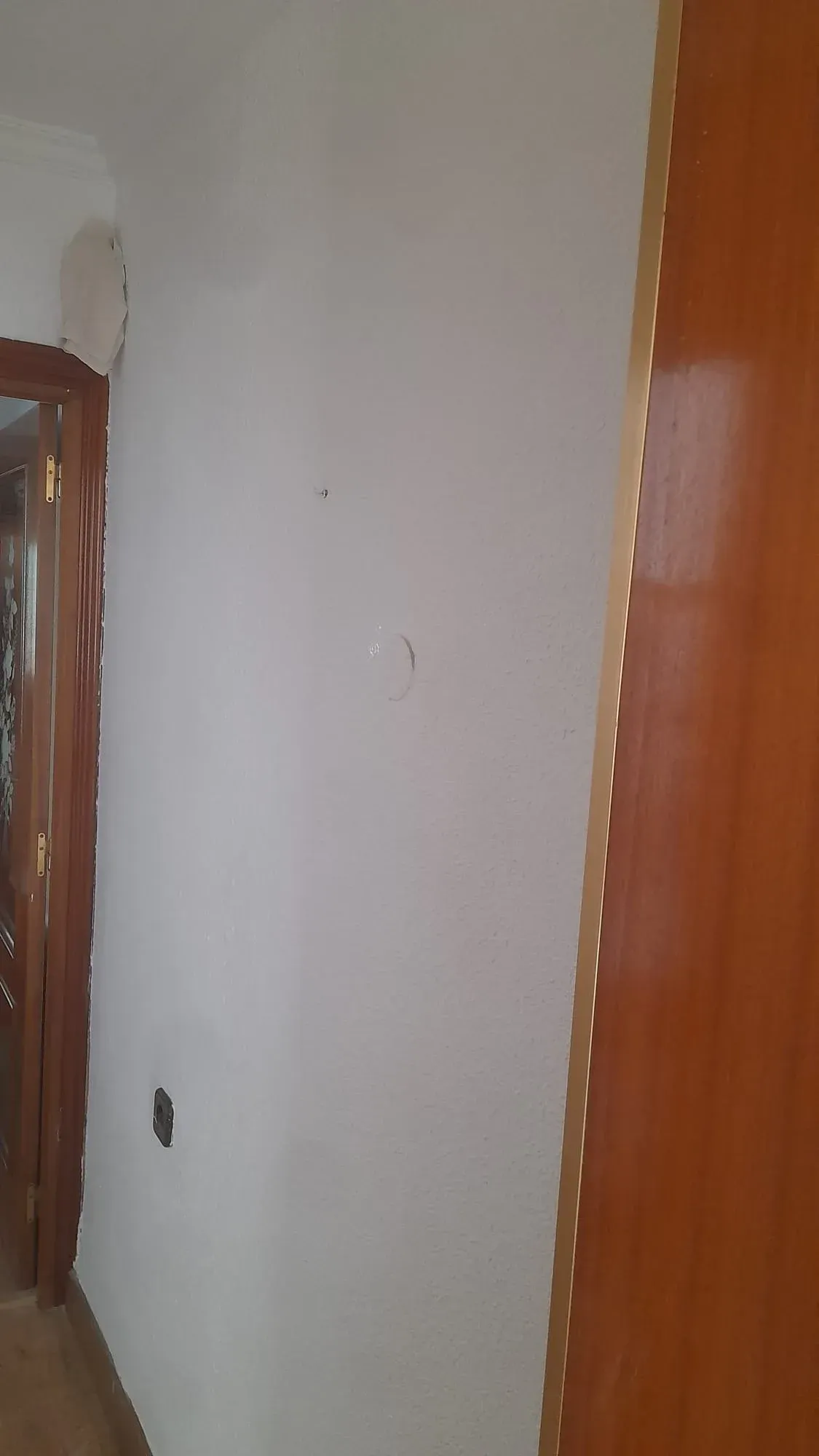 Una pared interior blanca junto a un marco de puerta de madera, con una pequeña marca en la pared cerca de la parte inferior.