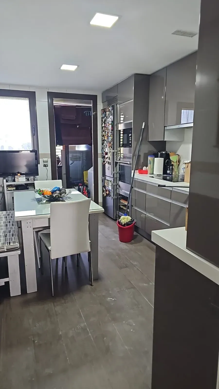 Cocina moderna con armarios grises, sillas de comedor blancas y una encimera abarrotada de objetos domésticos.