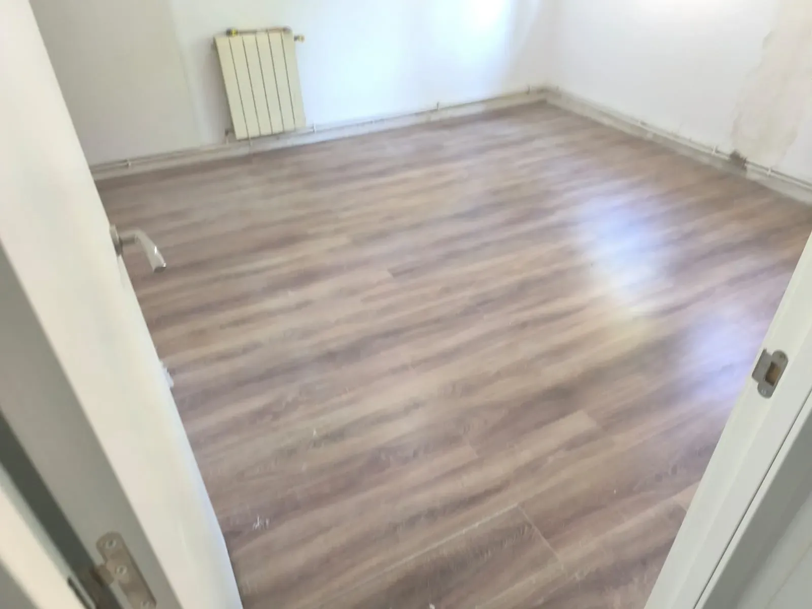 Habitación vacía con suelo de imitación madera, paredes blancas y un radiador junto a la pared del fondo.