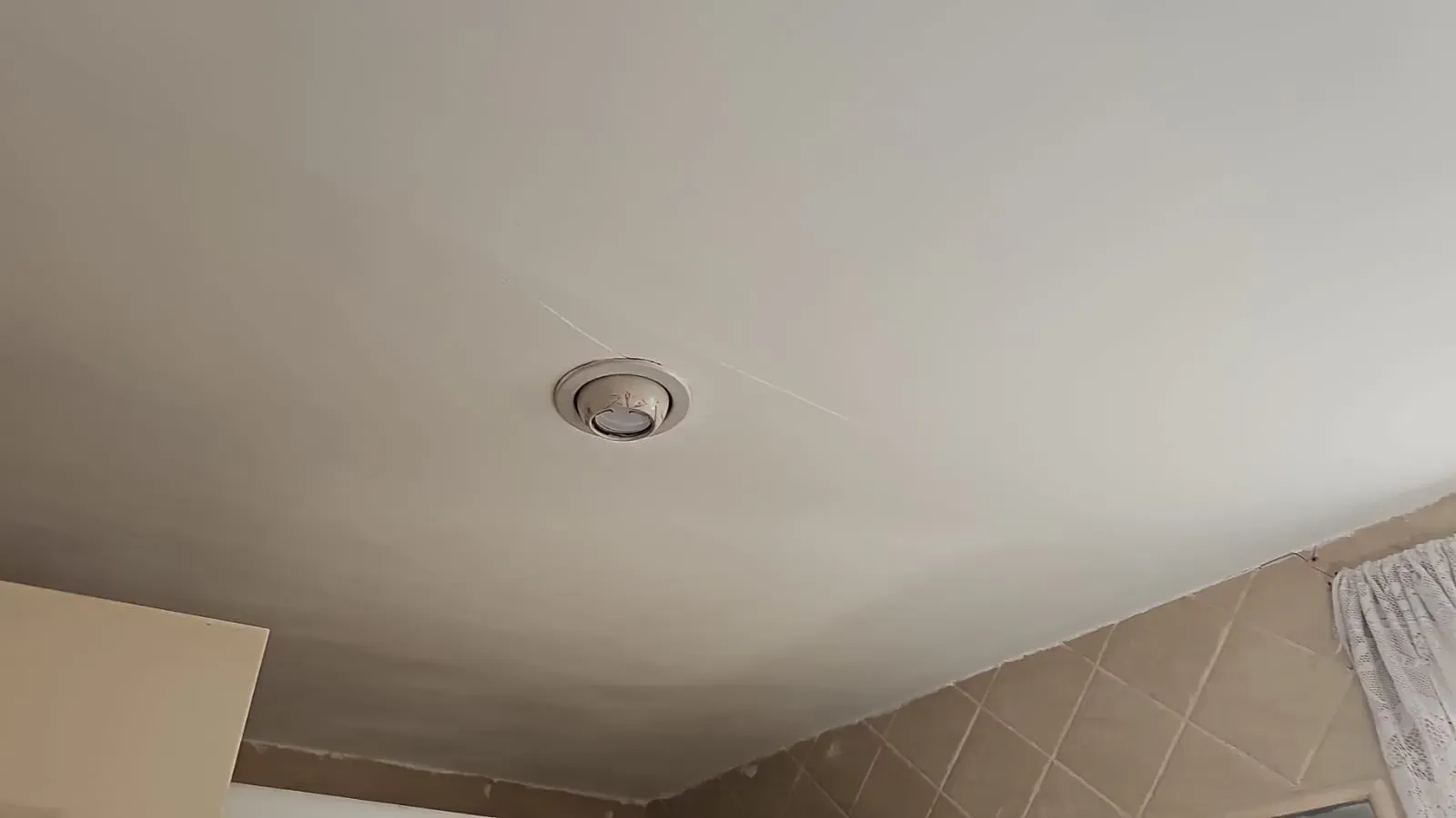 Techo con una lámpara redonda empotrada sobre una pared alicatada y una cortina.