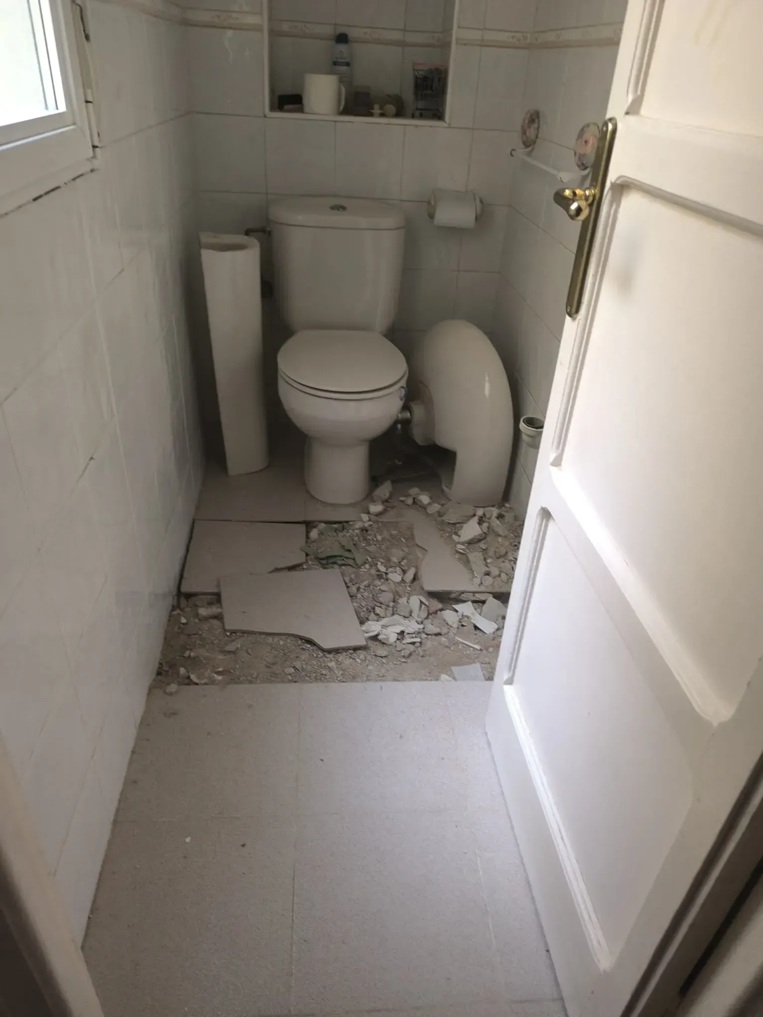 Baño pequeño dañado, con baldosas rotas en el suelo y un inodoro en el centro.