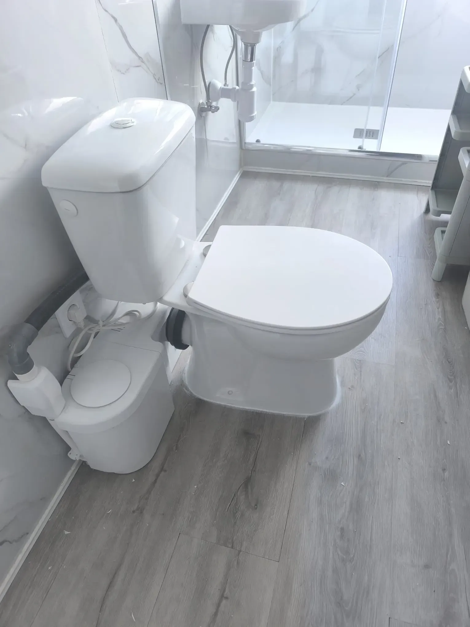 Baño pequeño de color blanco con inodoro, lavabo y ducha sobre suelo de imitación madera gris.