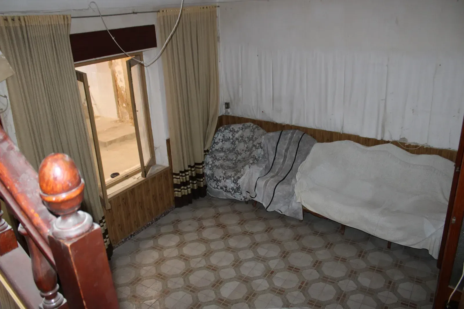Habitación pequeña con cama, alfombra estampada y ventana abierta con cortinas.
