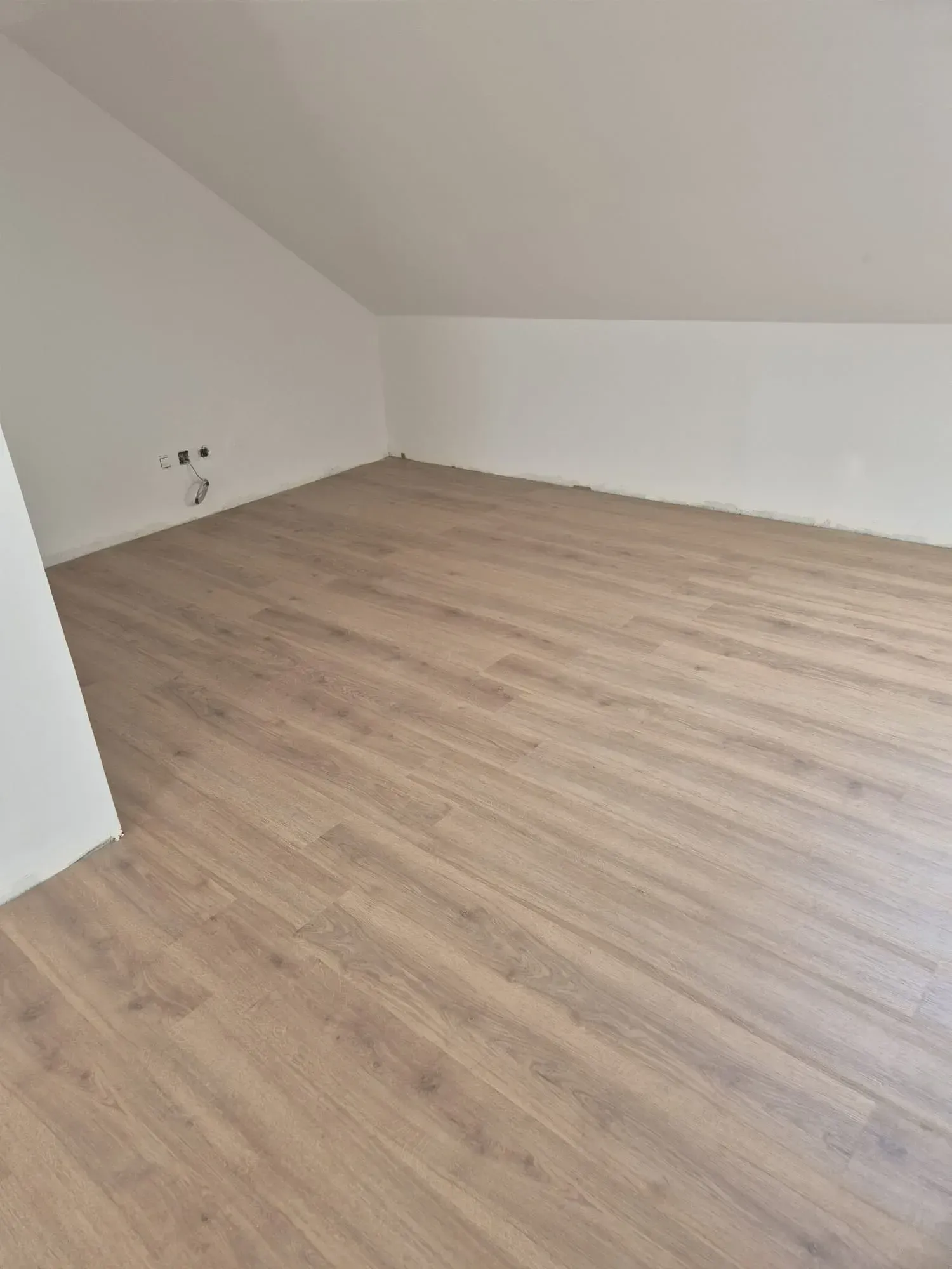 Habitación vacía con suelo de madera clara, paredes blancas y techo inclinado.