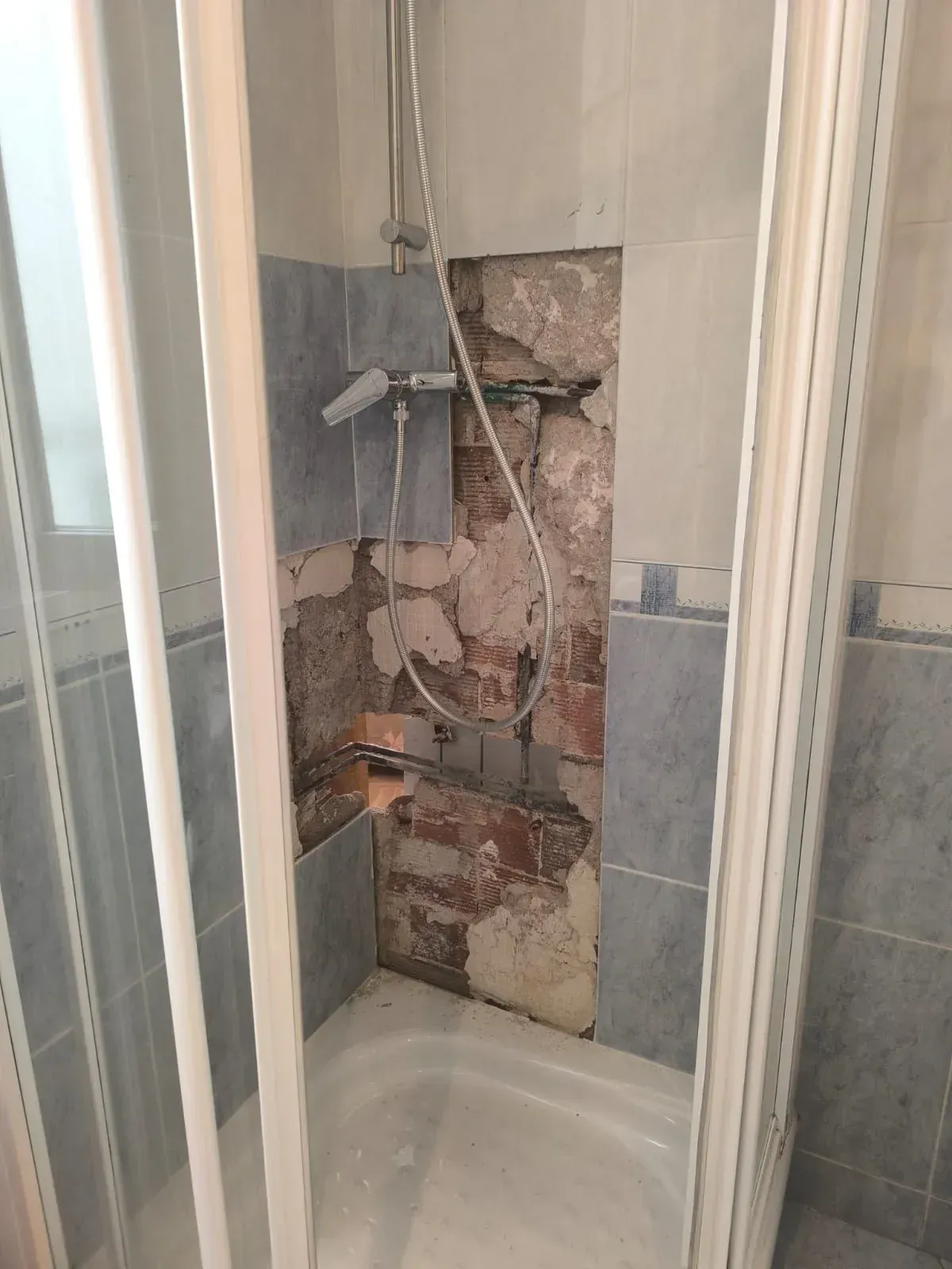 Pared de ducha dañada con tuberías expuestas y azulejos rotos en un pequeño baño.