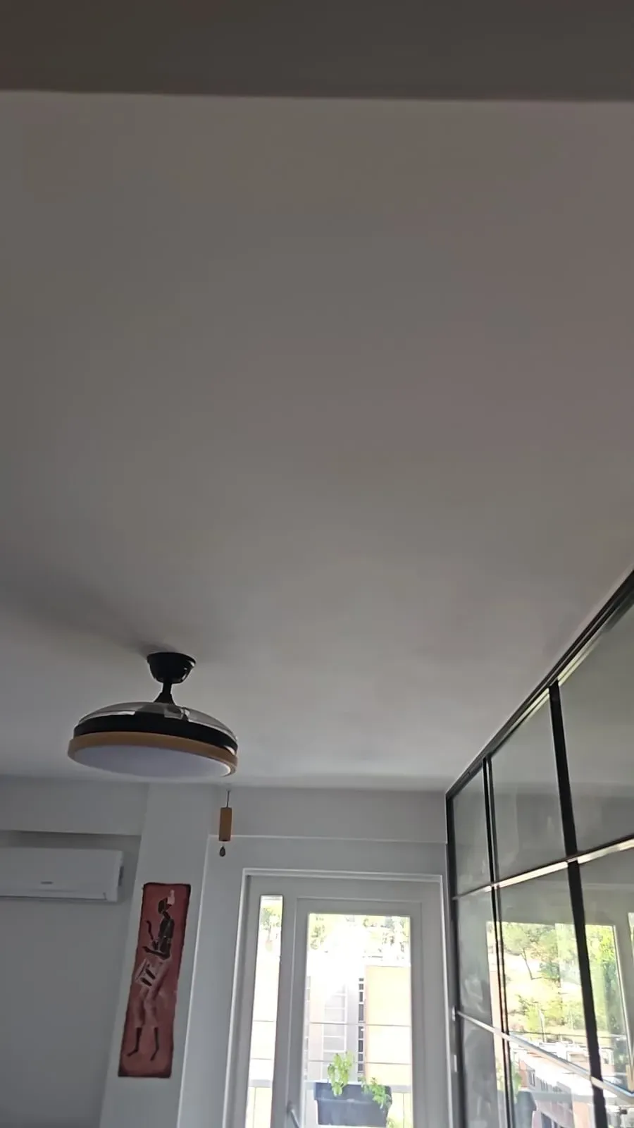 Pasillo interior con techo blanco, ventilador de techo, pared de cristal y puerta abierta a una zona exterior luminosa.