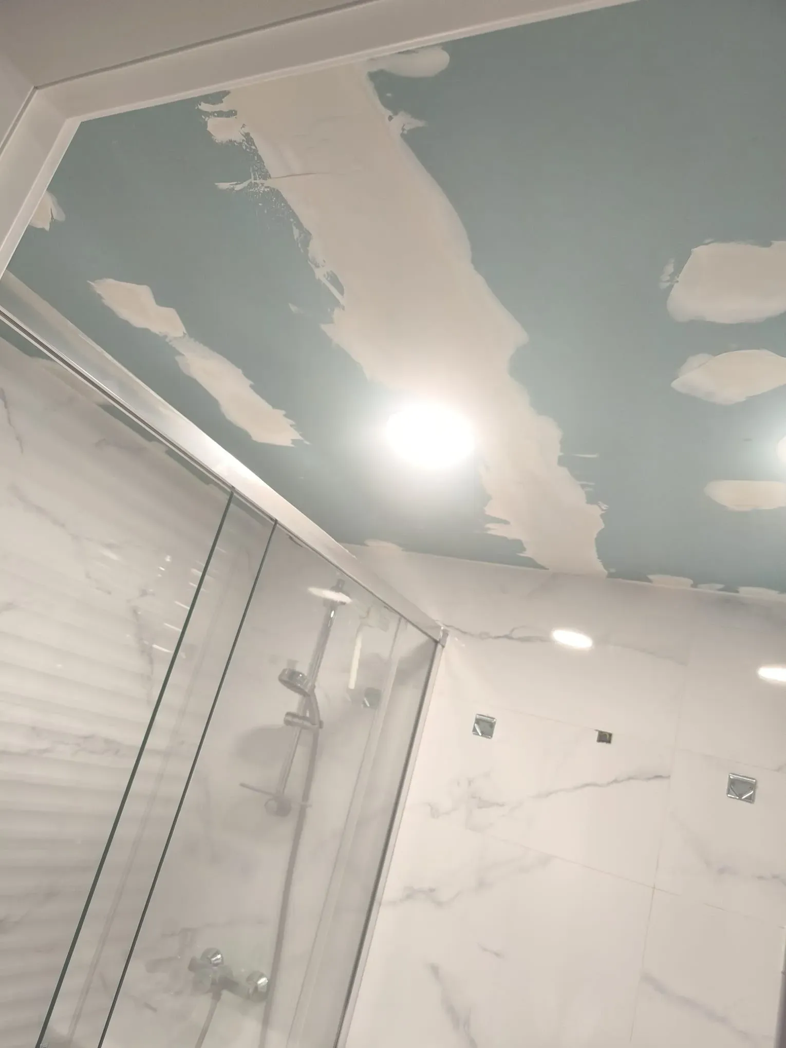 Ducha en el baño con puerta de cristal, paredes con aspecto de mármol y techo azul con manchas blancas que simulan nubes.
