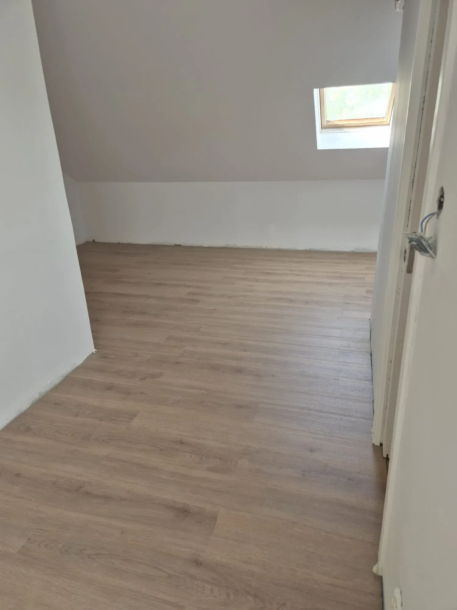 Habitación vacía con suelo de madera clara, paredes blancas y una pequeña claraboya.