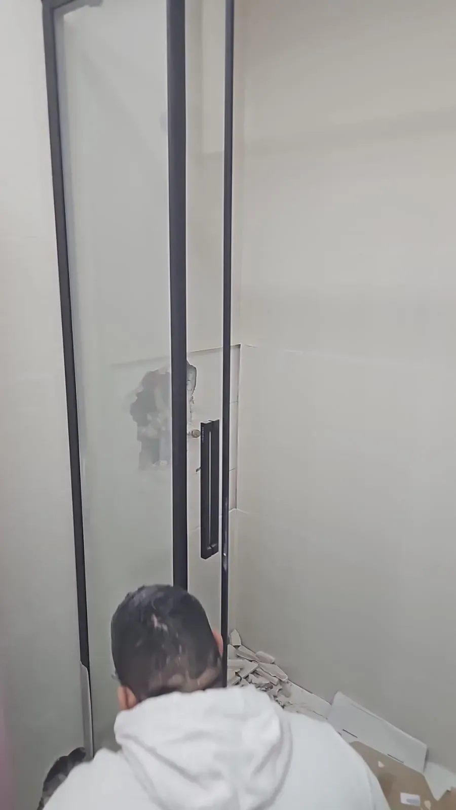 Hombre con sudadera blanca trabajando en una pared dañada junto al marco de una puerta de cristal.