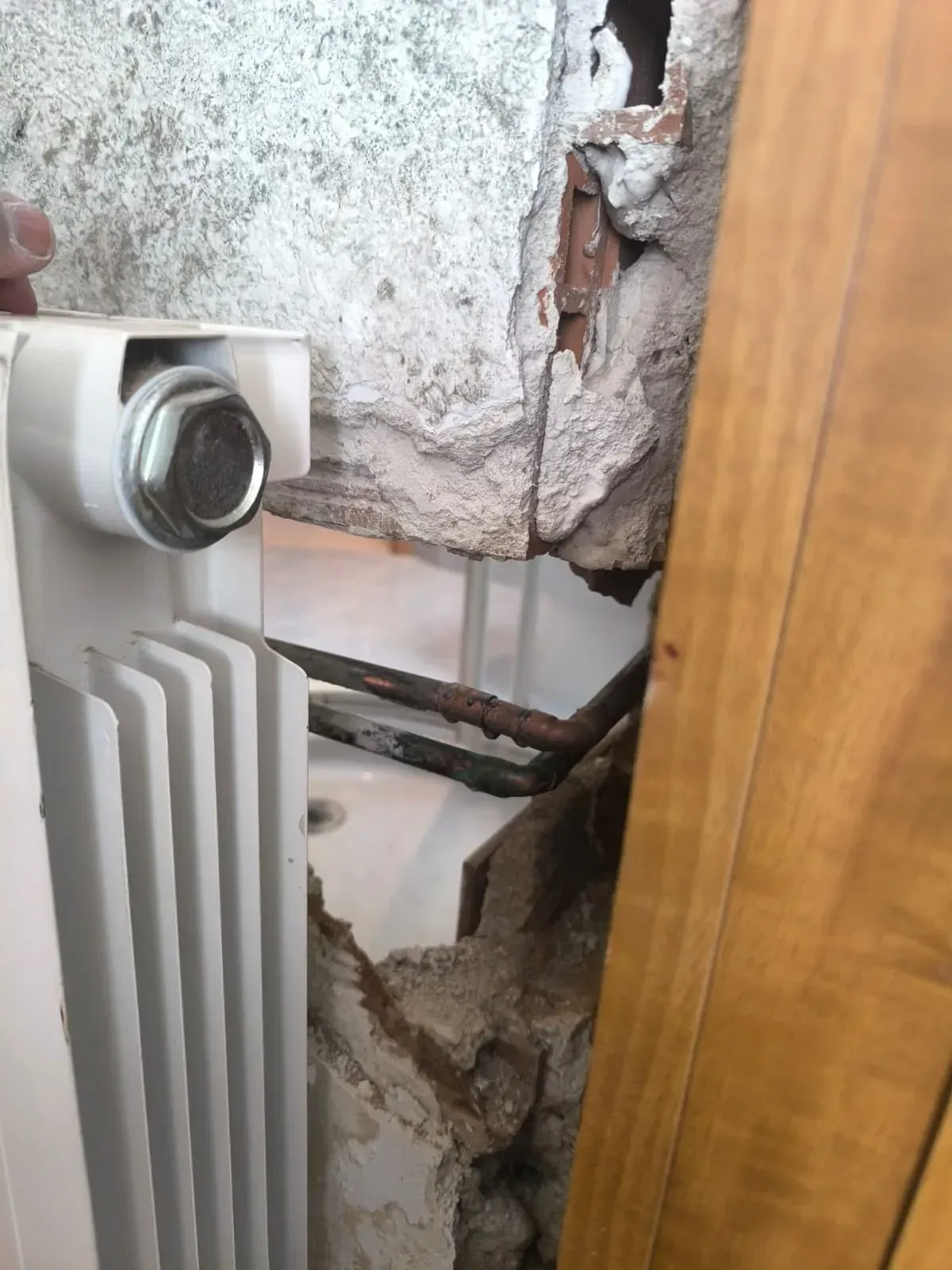 Pared dañada detrás de un radiador, con yeso desmoronándose y material estructural expuesto cerca de una puerta de madera.