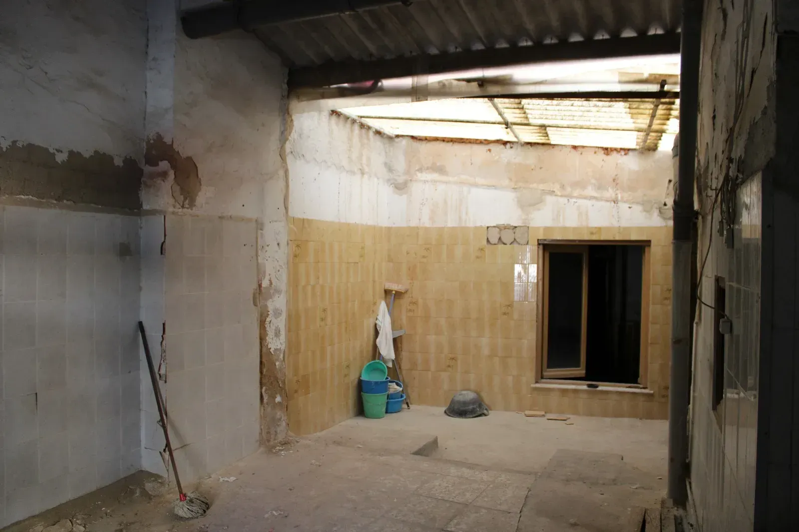 Patio en ruinas con paredes desconchadas, una abertura para una ventana y una escoba apoyada junto a bolsas apiladas.