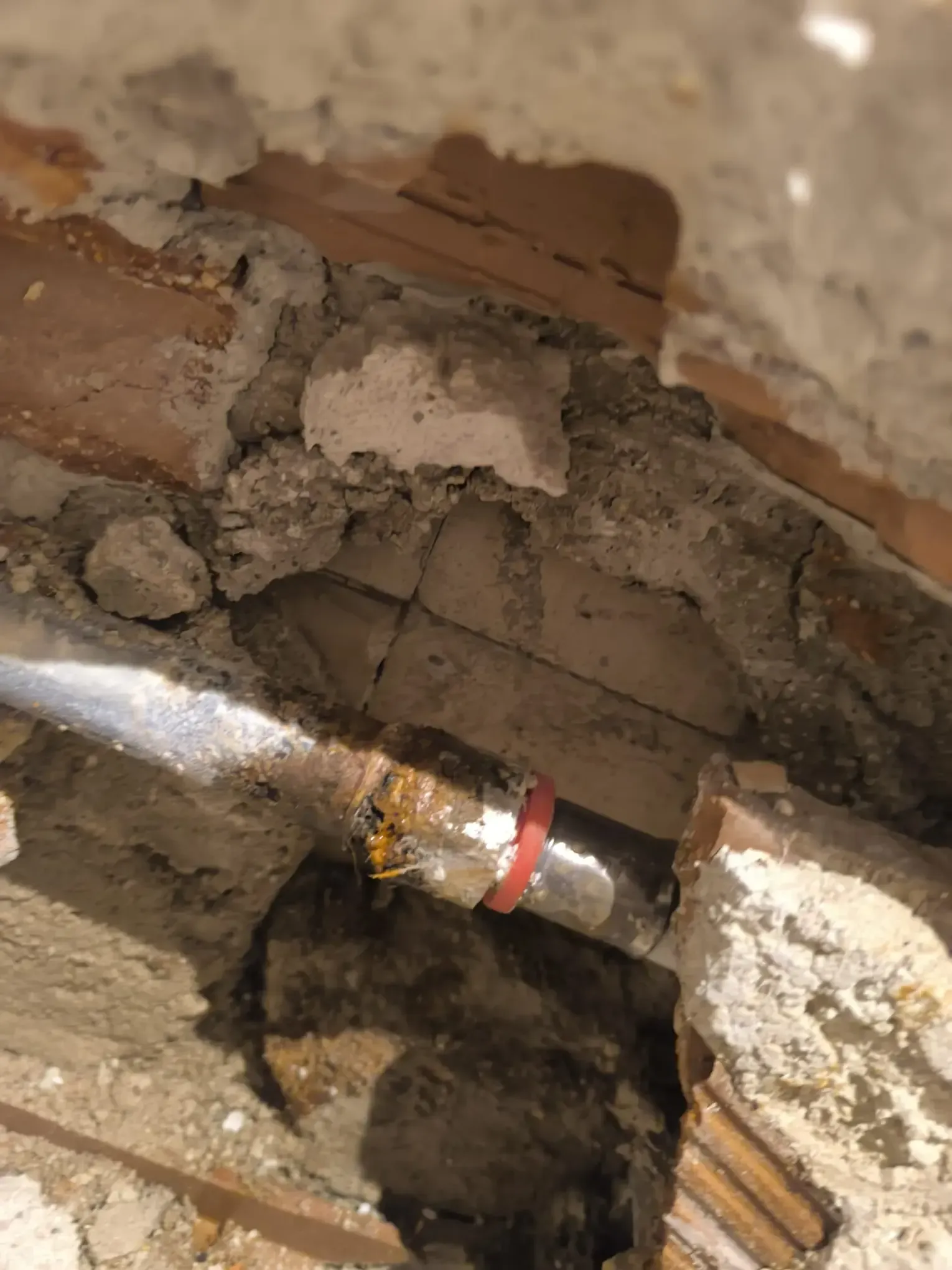 Tubería expuesta en una cavidad de pared dañada con yeso desmoronado y una banda de acoplamiento roja.