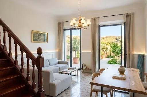 Rincón de comedor luminoso con escalera, sofá blanco y puertas que dan a un patio ajardinado soleado.