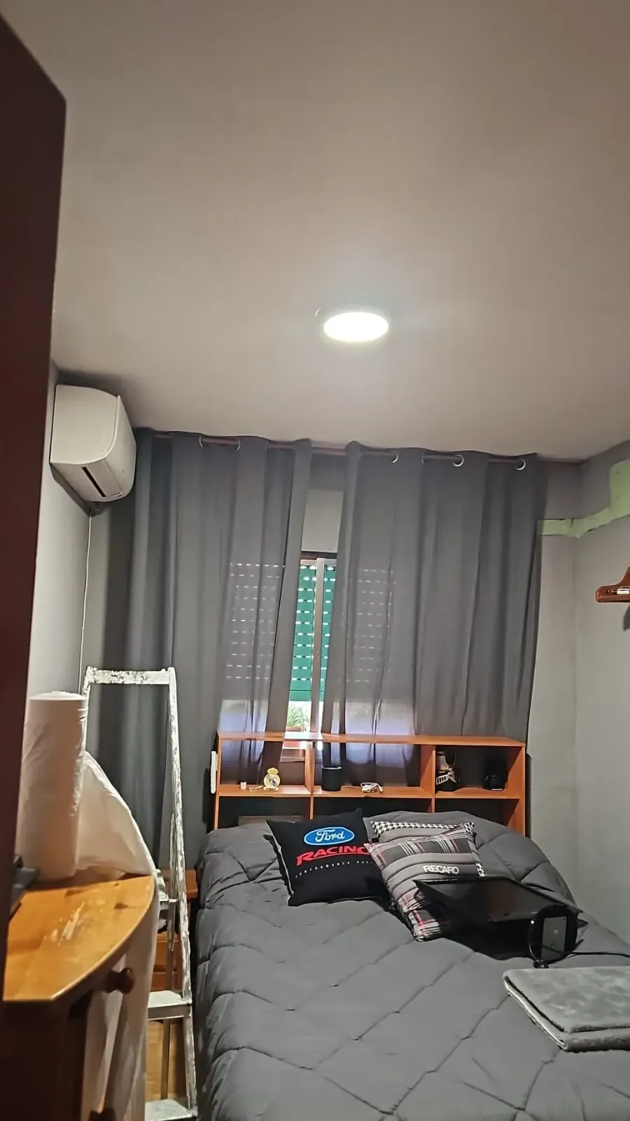 Habitación pequeña con una cama, cortinas grises, escritorio y lámpara de techo cerca de una ventana.