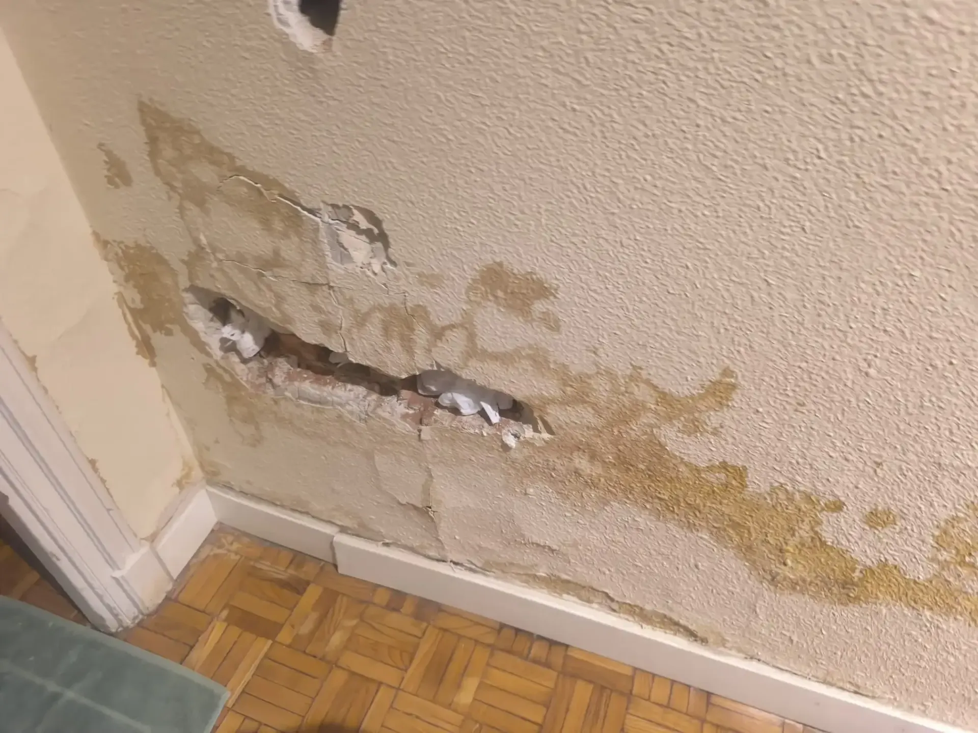 Pared dañada con pintura descascarada y yeso expuesto sobre un piso y rodapié de madera.