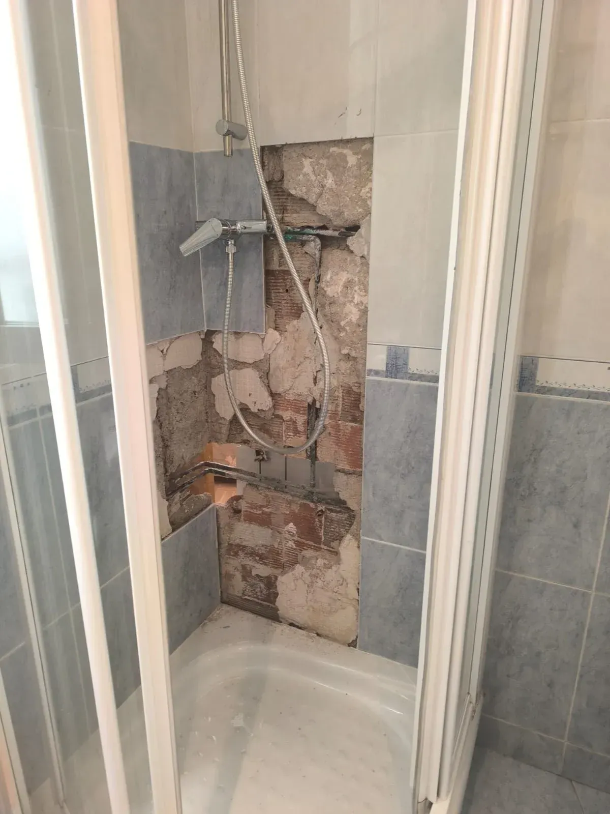 Cabina de ducha con tuberías a la vista y pared dañada detrás de los azulejos.