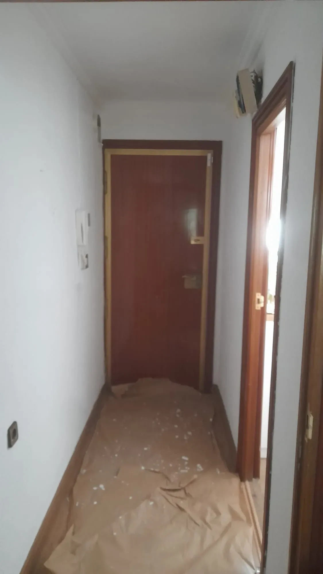 Pasillo estrecho con paredes blancas, una puerta de color marrón rojizo y un suelo cubierto con papel protector
