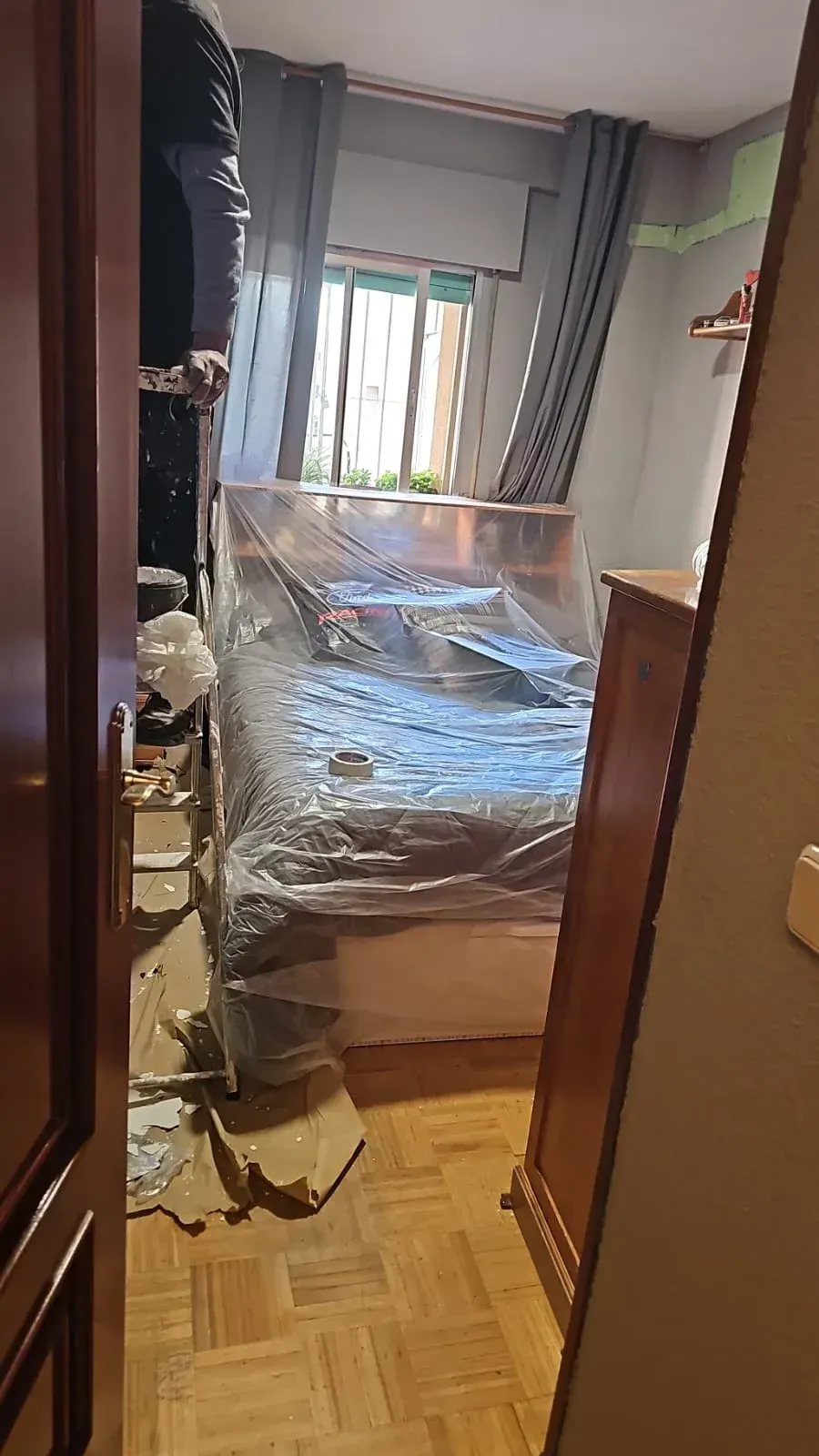 Habitación estrecha con una cama envuelta en plástico, cortinas grises y una ventana al fondo.