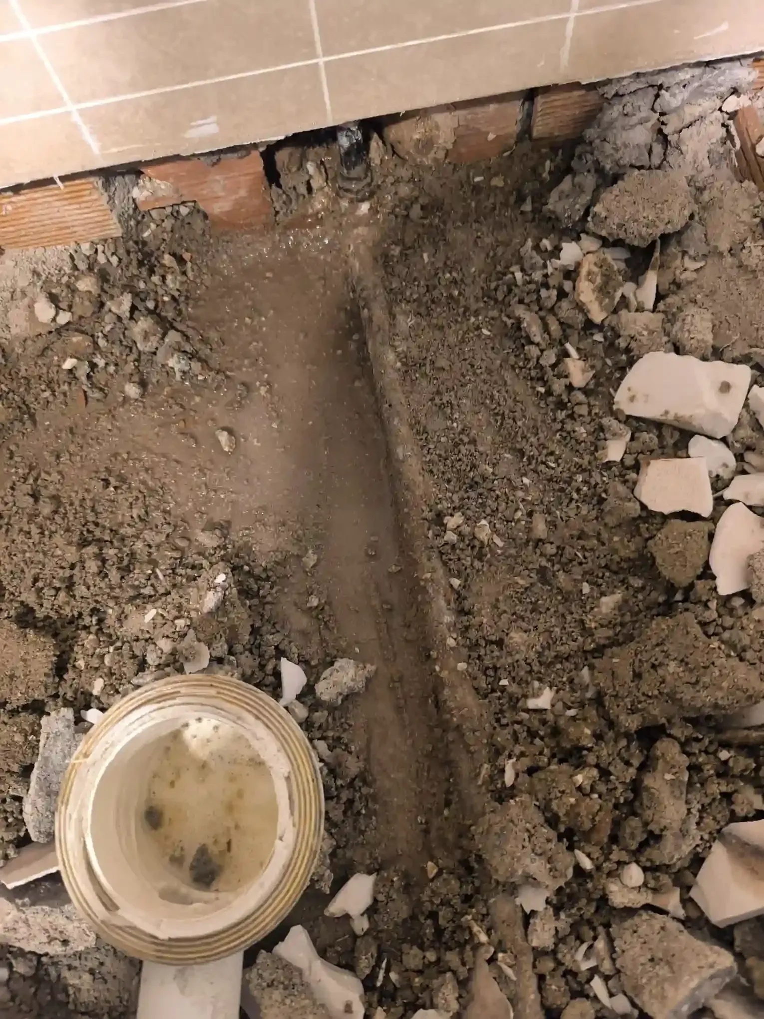 Suciedad y escombros expuestos alrededor de la abertura de una tubería de desagüe en la base de una pared alicatada.