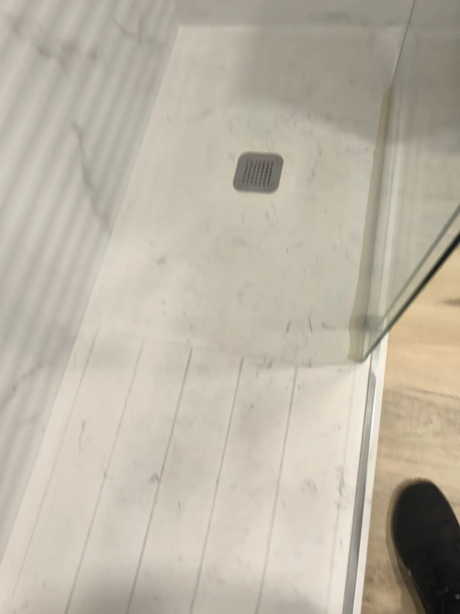 Suelo de ducha con azulejos blancos, desagüe cuadrado y parte de un zapato negro visible en el borde.