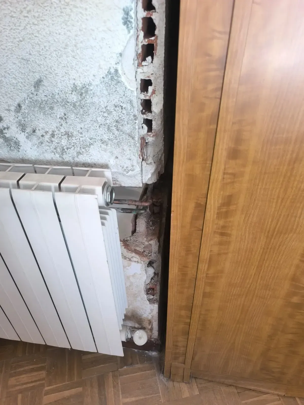 Pared dañada por un radiador y un marco de puerta de madera, con ladrillos a la vista y yeso desconchado.