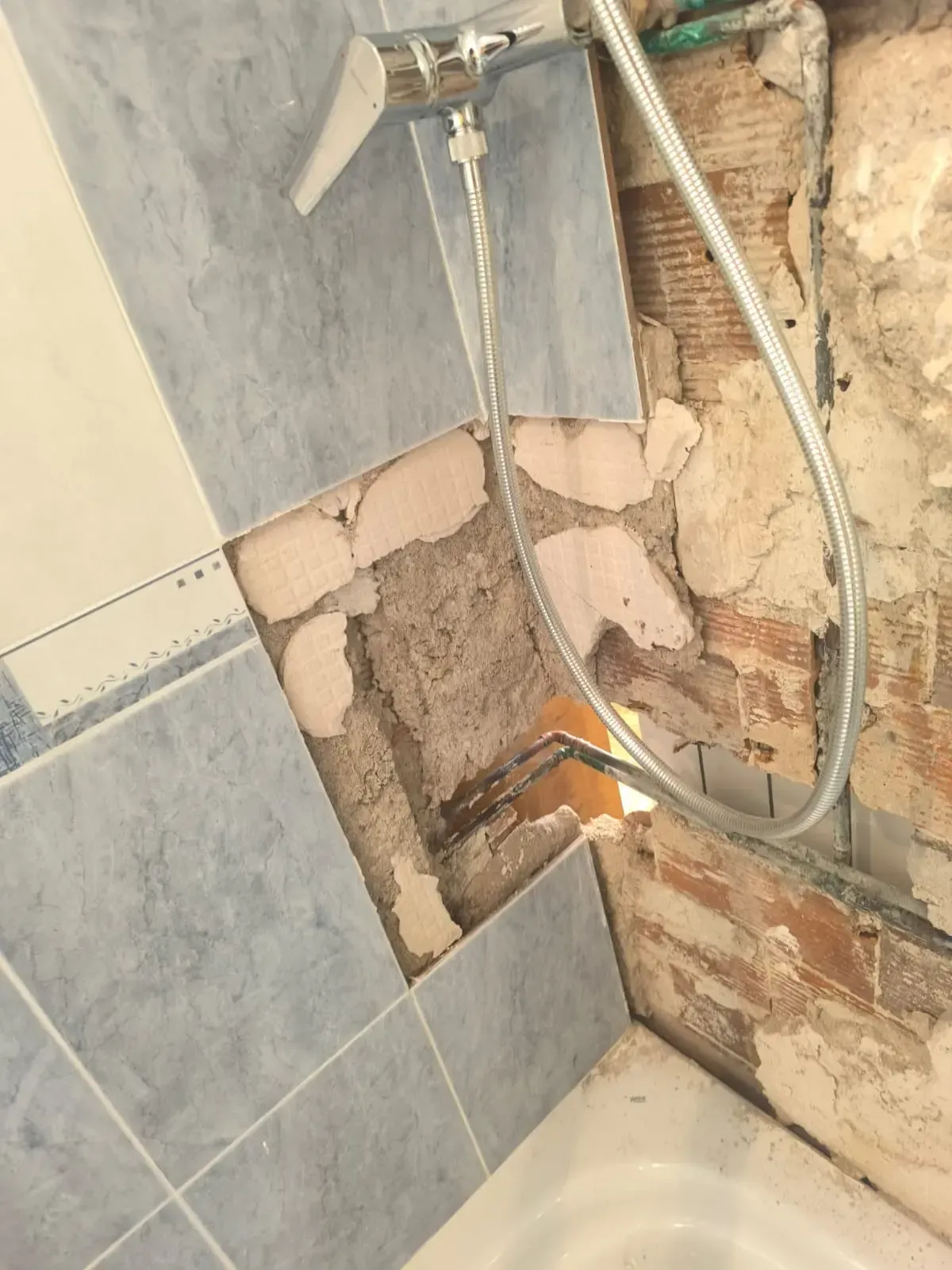 Pared de la ducha dañada con tuberías expuestas, azulejos agrietados y ladrillos expuestos cerca de la bañera.