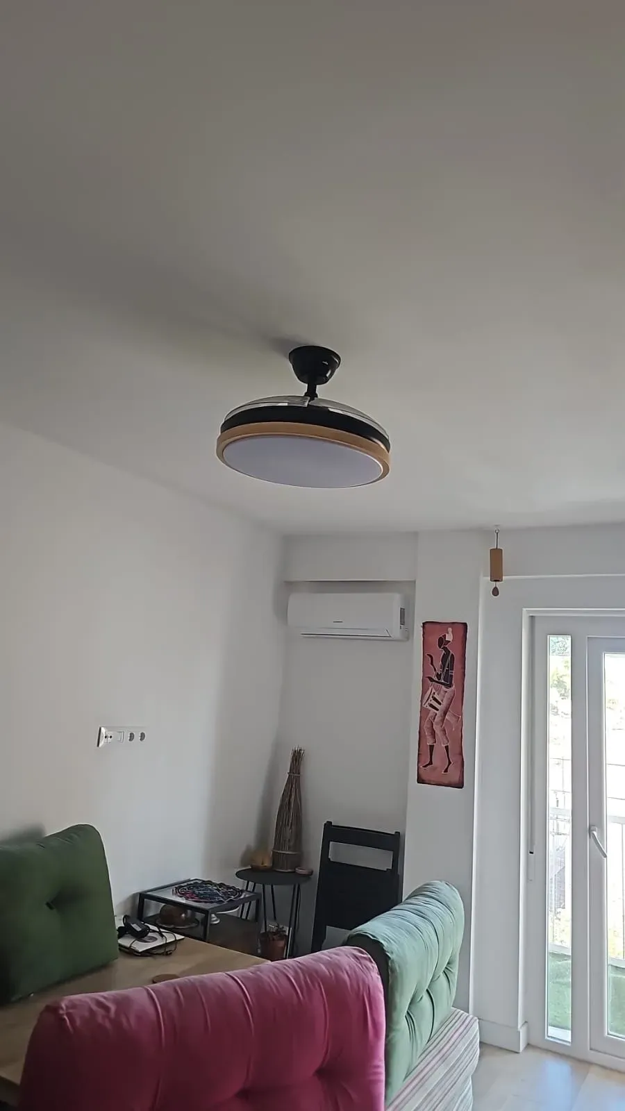 Sala de estar con sofá verde y rosa, lámpara de techo, aire acondicionado mural y puerta de cristal que da al balcón.